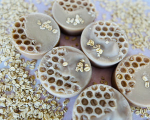 6 cream soap rounds sit on a bed dried oats. The bars themselves have a honeycomb texture and some ground oat on them.